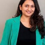 A woman with long dark hair, wearing a green blazer and a black shirt, smiles warmly against a plain background.