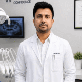 A dentist in a white coat stands in a modern dental office, with a dental chair and an X-ray image in the background.